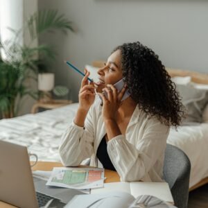 African American woman multitasking at home desk with laptop and phone, embodying modern remote work life.
